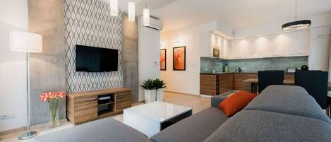 Una sala moderna con sofá, mesa de centro y televisor. Hay una zona de cocina con armarios y una mesa de comedor.