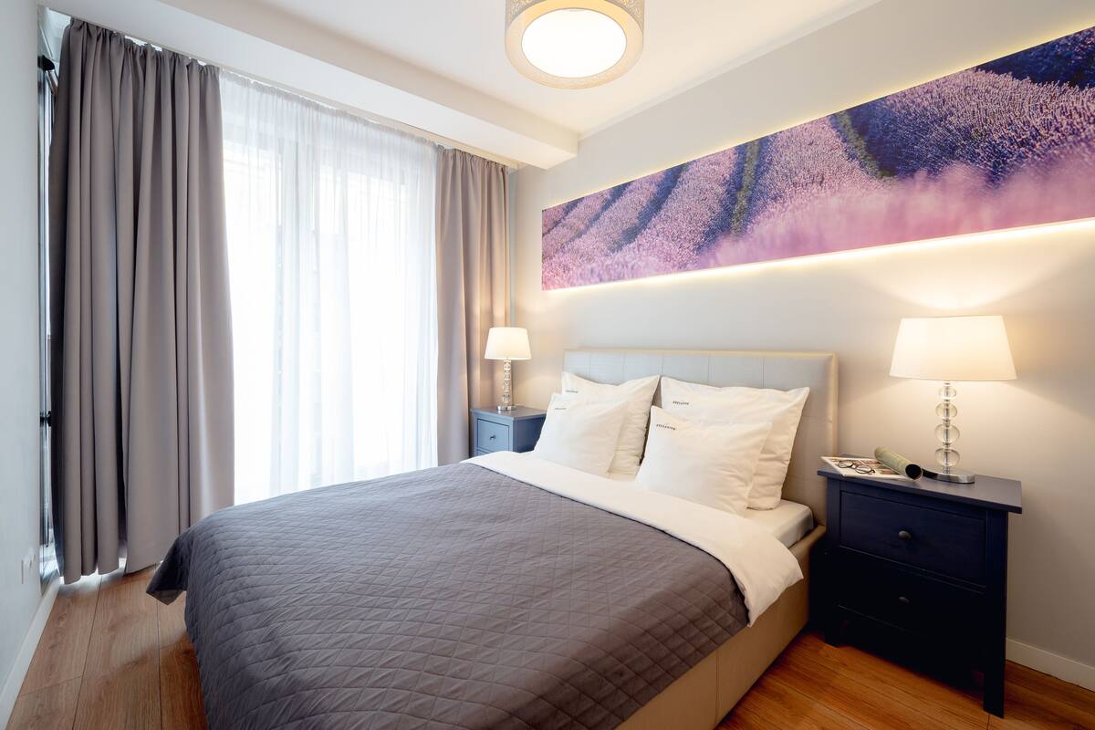 A neatly made bed with a quilted bedspread, two bedside lamps, a nightstand with a lamp, and a large abstract painting on the wall.
