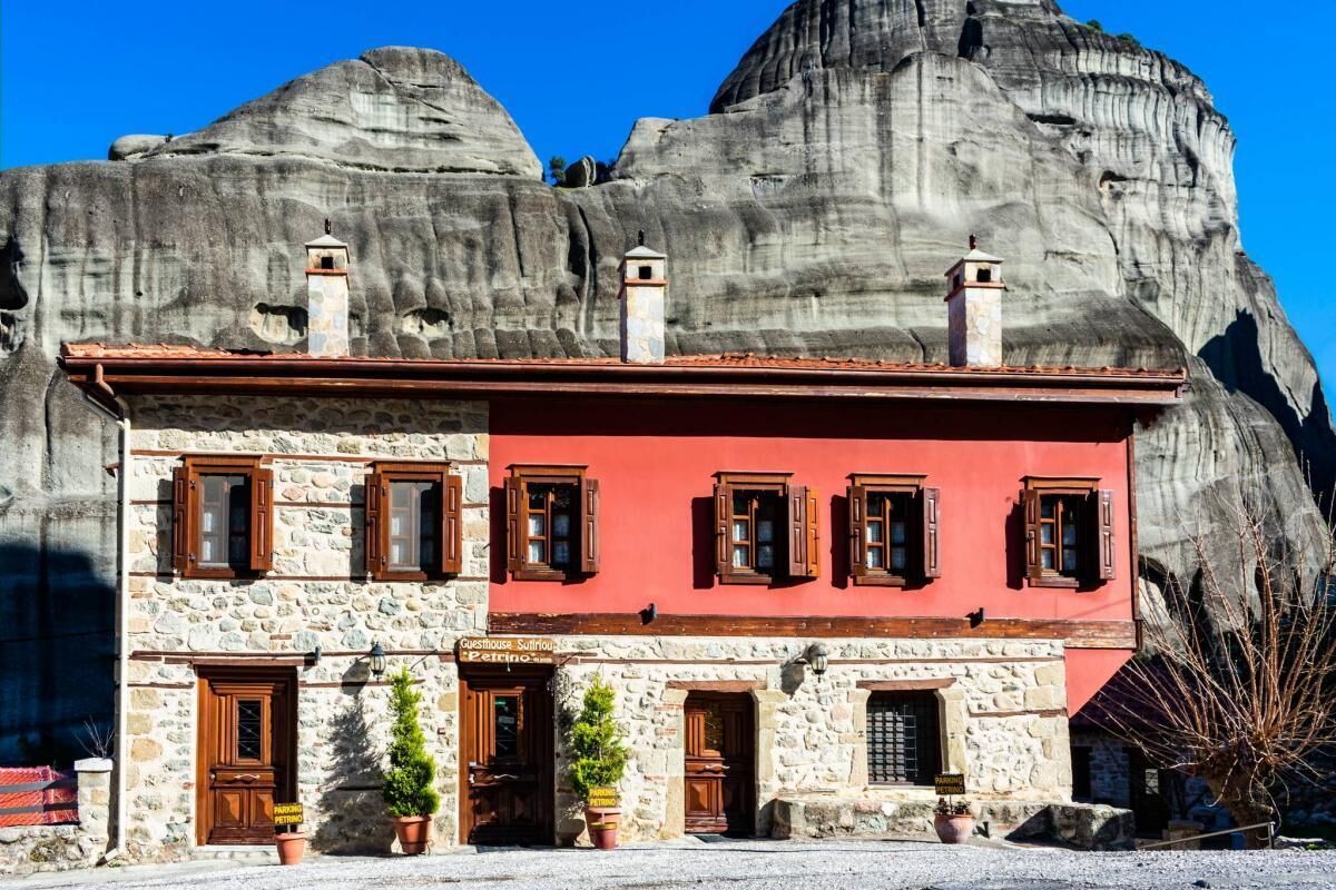 Un edificio de piedra con fachada roja y contraventanas de madera, situado frente a una gran formación rocosa.