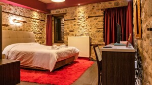 A bedroom with a stone wall, a bed with a red rug, a desk with a chair, and a mirror. - Guesthouse Sotiriou (Kalabaka)