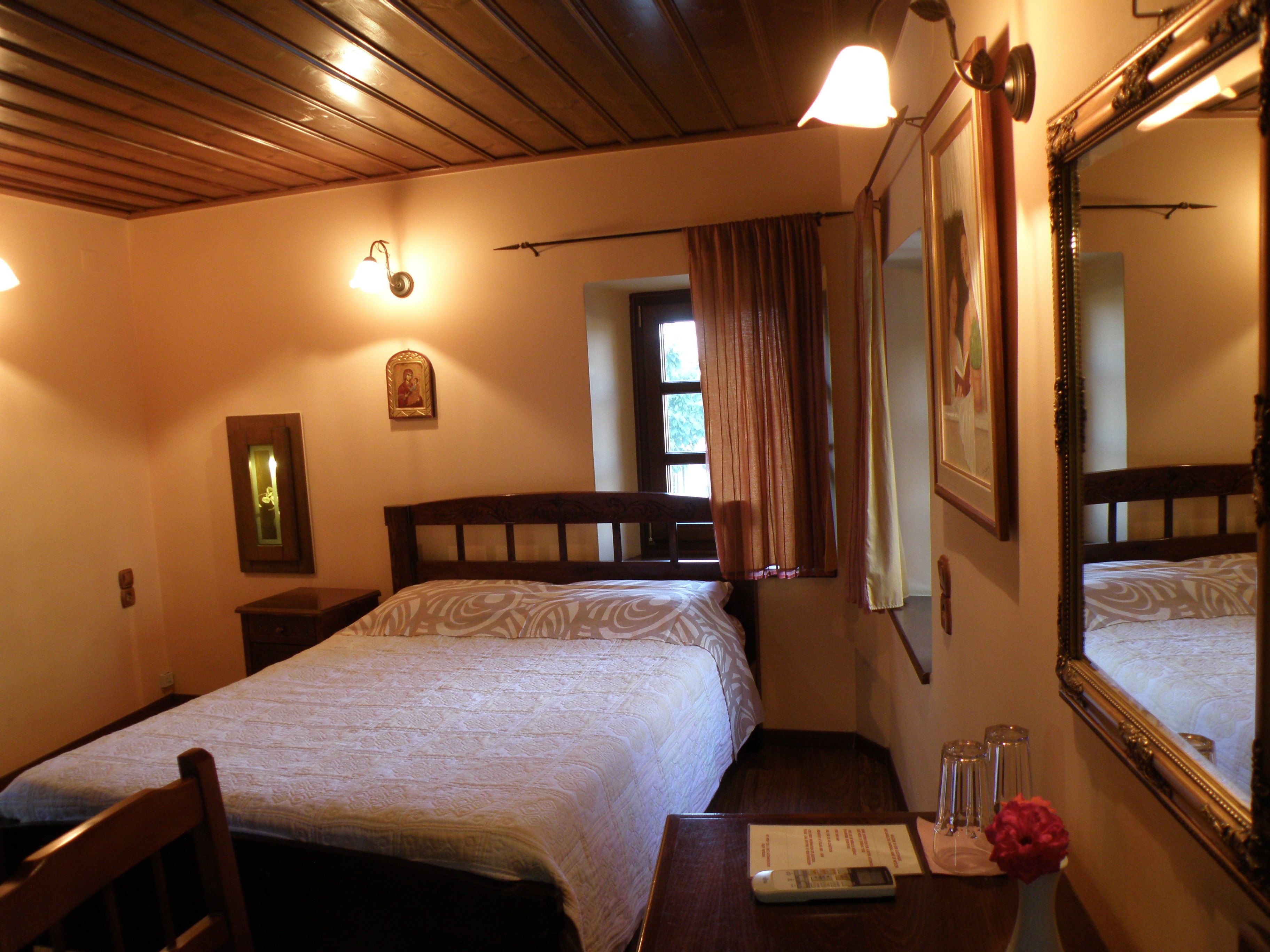 A hotel room with a bed, a nightstand, a mirror, a window with curtains, and a wall-mounted light fixture.