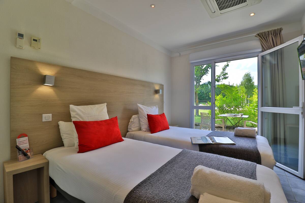Habitación de hotel con dos camas, cada una con ropa blanca y almohadas rojas, un cabecero de madera y vistas al exterior a través de un ventanal corredera.