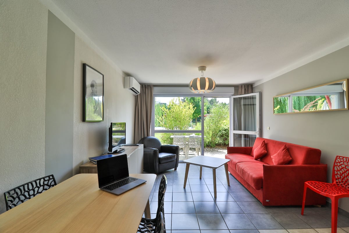 Un salón moderno con un sofá rojo, una silla negra, una mesa de madera con un ordenador portátil y vistas a la vegetación a través de grandes ventanales.