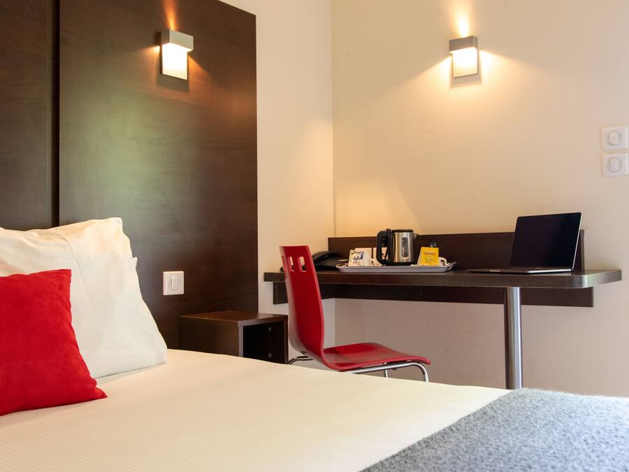 A hotel room with a bed, a desk with a laptop, a red chair, and two wall-mounted lights.