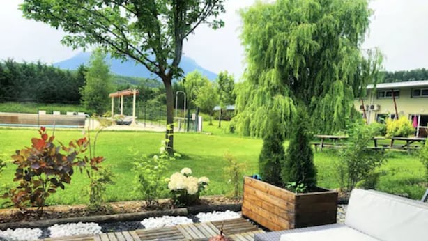 Eine Terrassenfläche mit einer Couch, einem kleinen Tisch und einer Zierlaterne. Ein Garten mit Baum und Blumen sowie eine hölzerne Bank sind ebenfalls vorhanden.