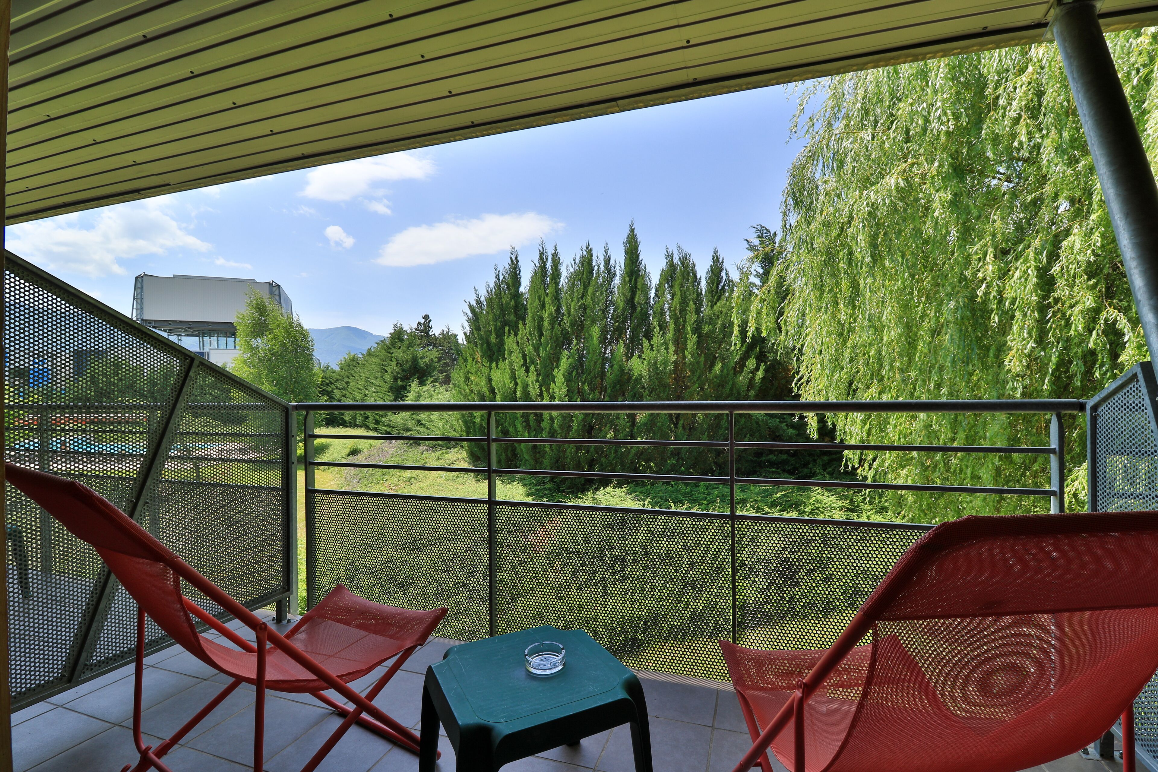 A balcony with two red chairs and a small table, overlooking a forested area with a clear sky.