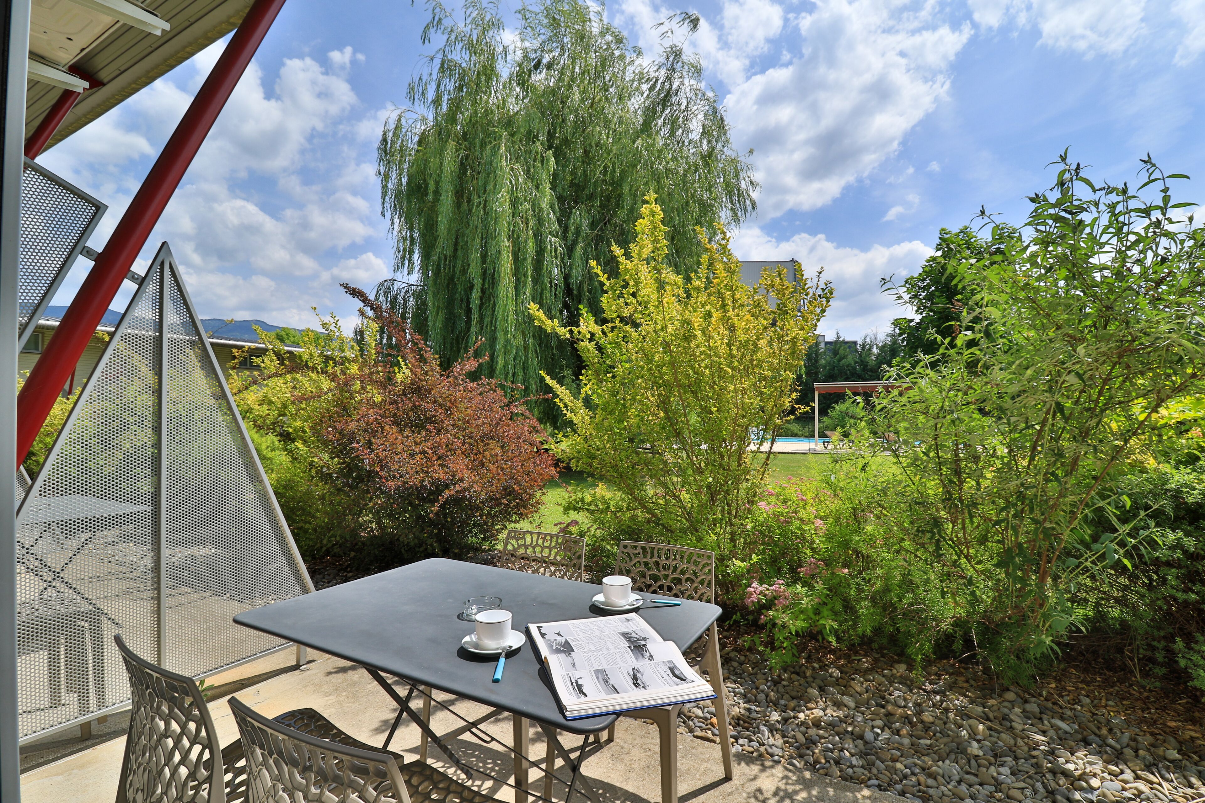 An outdoor seating area with a table and chairs, surrounded by greenery and a view of a building in the background.