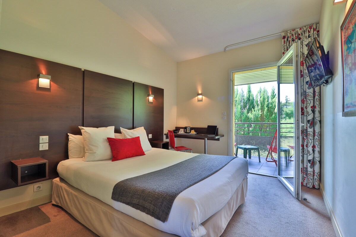 A modern hotel room with a large bed, a desk, and a balcony with outdoor seating.