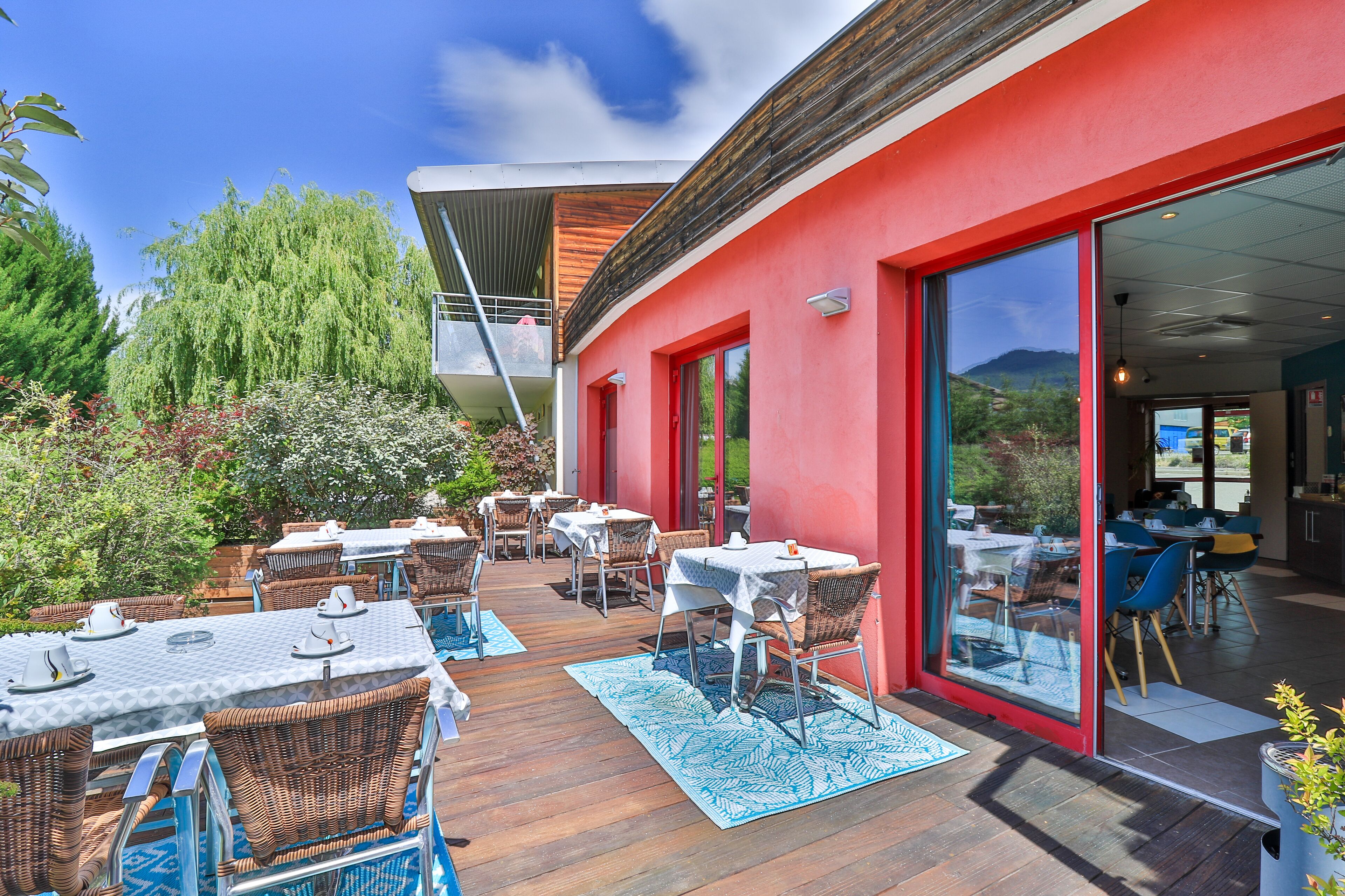 Un patio al aire libre con mesas y sillas, un edificio rojo con grandes puertas de vidrio y vistas a las montañas.