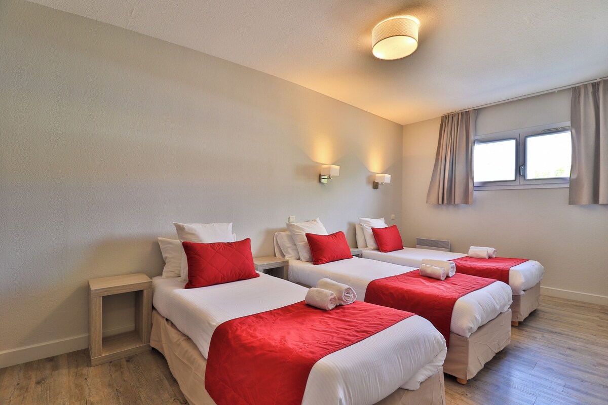 A hotel room with three beds, each with white bedding and red accents, a small bedside table, a wall-mounted light fixture, and a window with curtains.