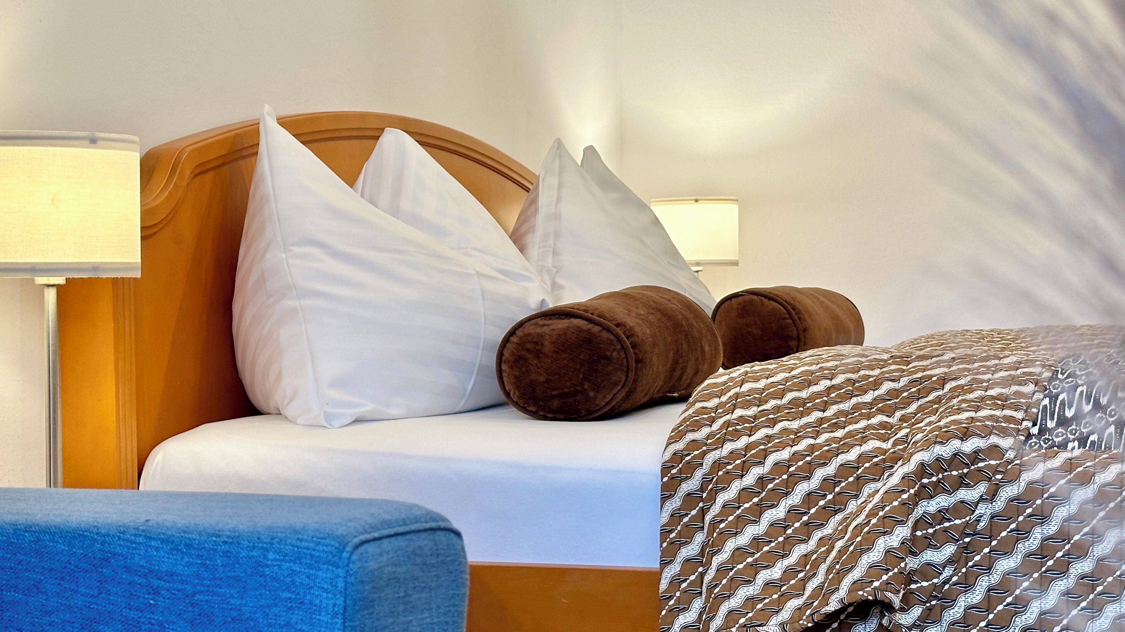 Ein ordentlich bezogenes Bett mit weißen und braunen Kissen, einer gemusterten Decke und einem blauen Stuhl.