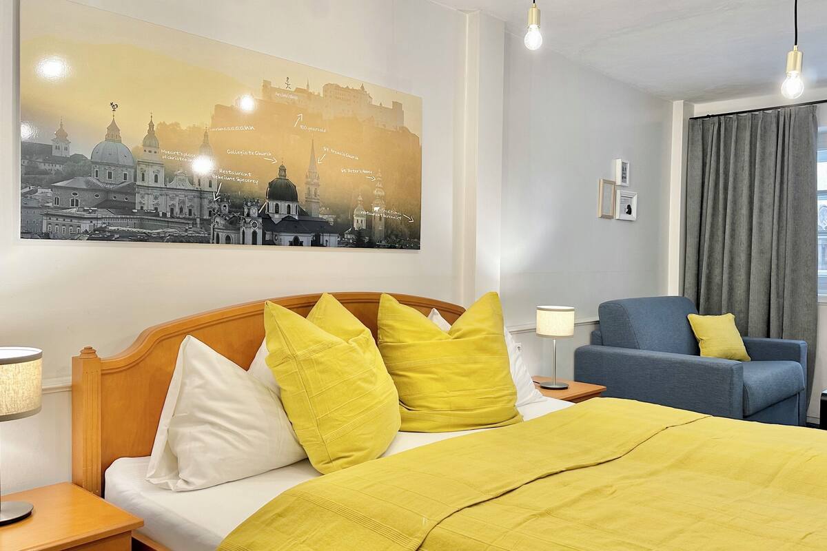 A hotel room with a bed, a chair, a lamp, and a large cityscape painting on the wall.