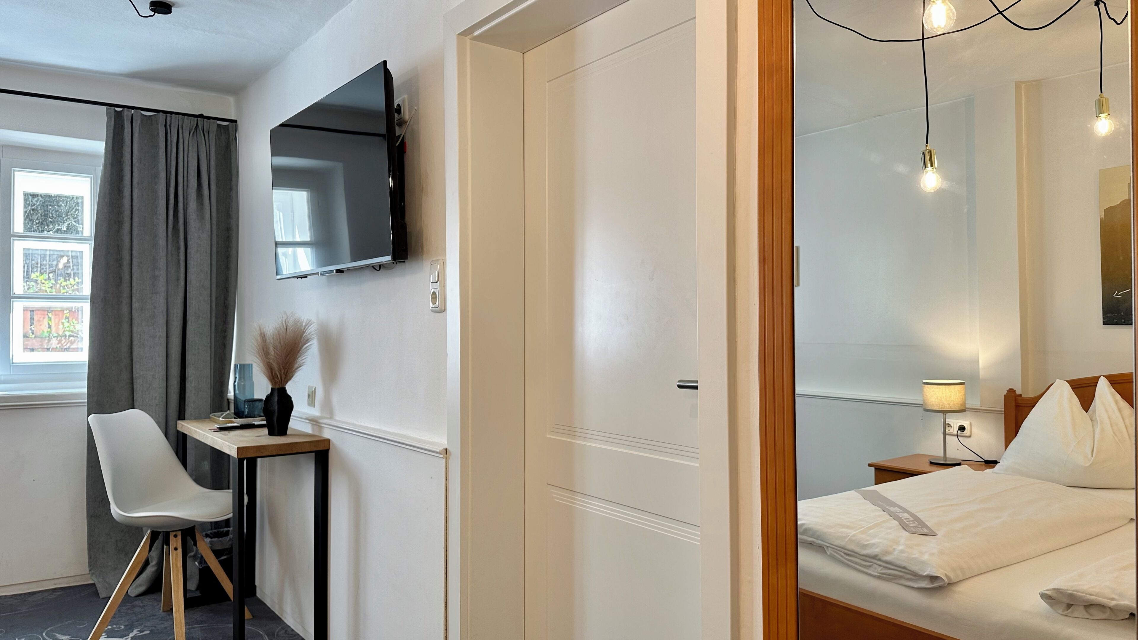 Una camera d'albergo con un letto, una scrivania con una sedia, una TV a schermo piatto fissata al muro e una finestra con tende.