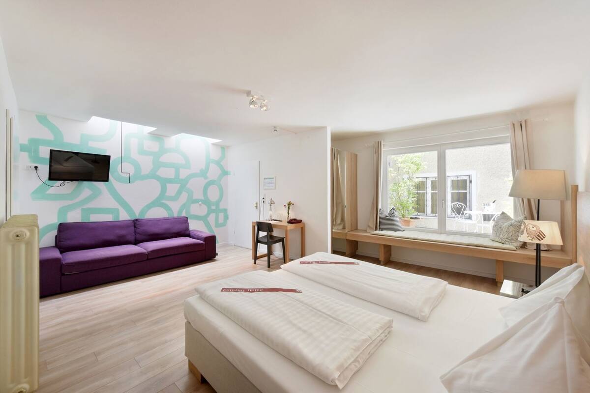 A modern hotel room with a bed, a purple sofa, a desk, a TV mounted on the wall, and a window with a view.