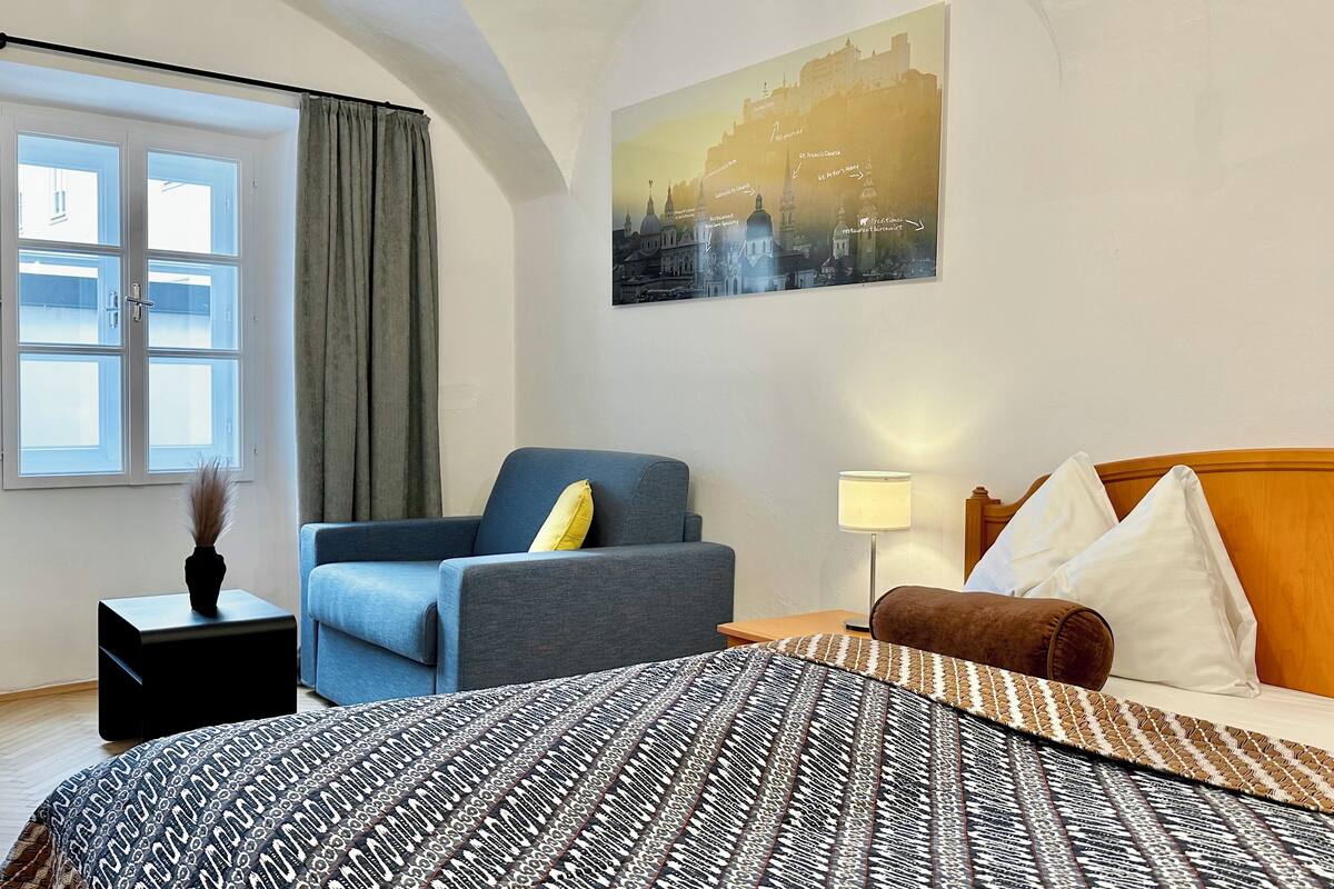 A hotel room with a bed, a blue armchair, a wooden headboard, a nightstand, and a painting of a cityscape.