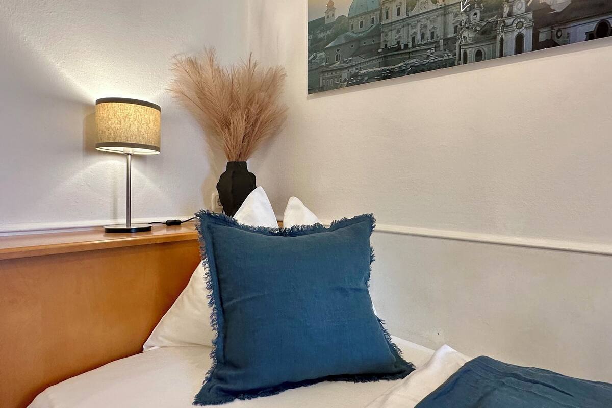 A neatly made bed with a blue pillow, a lamp, and a vase with dried plants on a wooden nightstand.