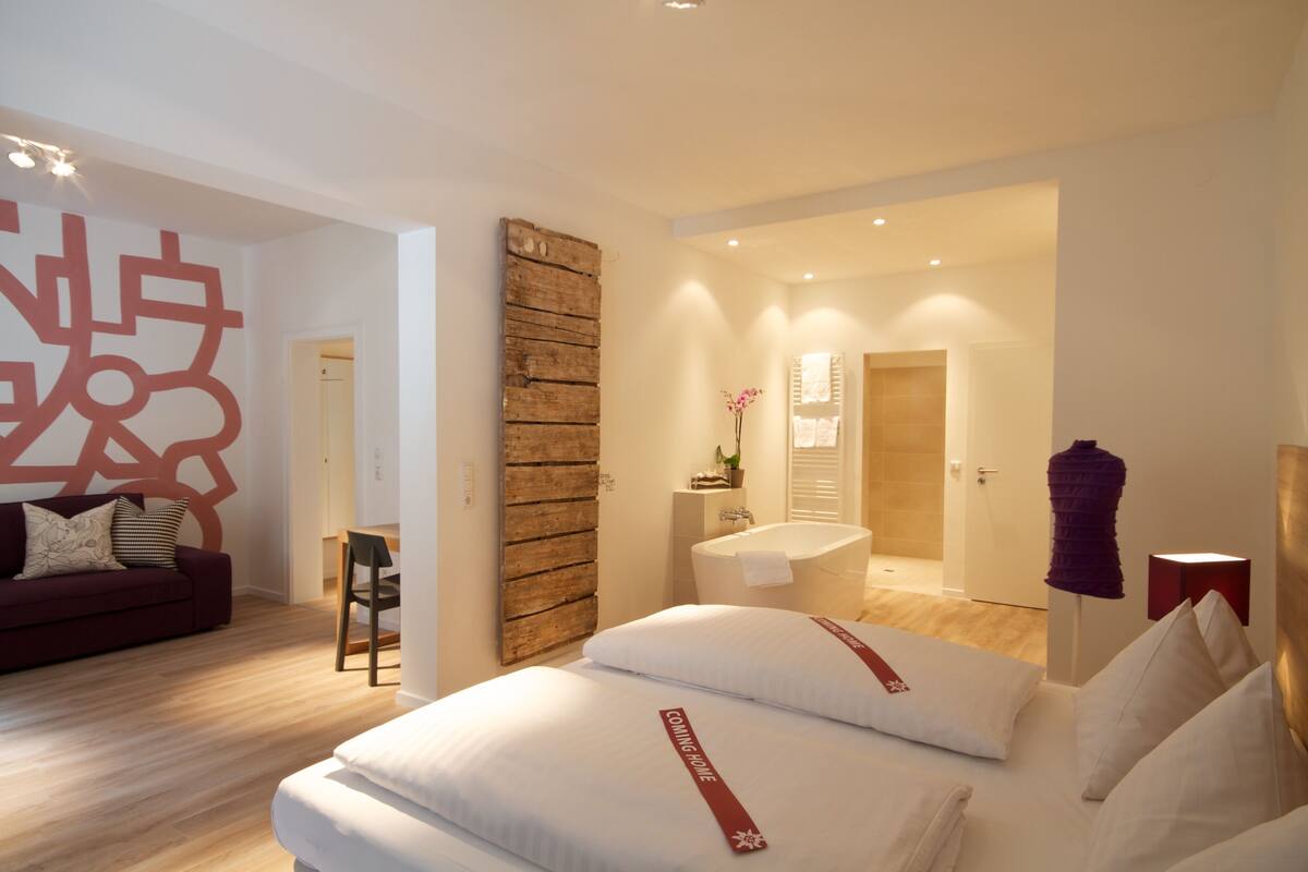 A modern hotel room with a large bed, a wooden accent wall, a bathroom with a bathtub, and a living area with a sofa and a decorative wall piece.