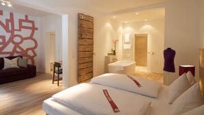 Ett modernt hotellrum med en stor säng, en vägg med träpanel som accent, ett badrum med badkar och ett vardagsrum med en soffa och ett dekorativt väggobjekt.
