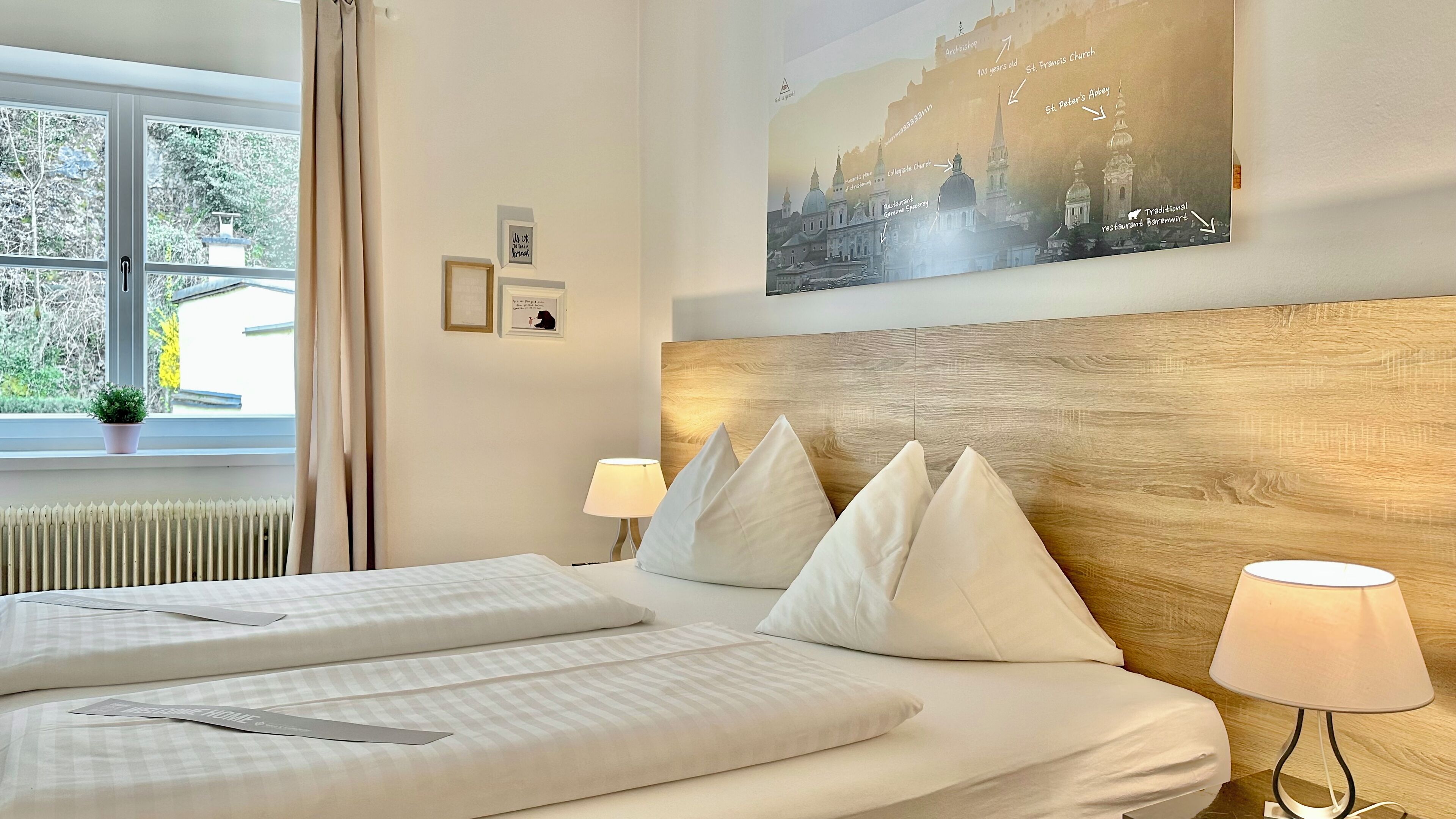 A hotel room with a large bed, two bedside tables, a window with curtains, a wall-mounted painting, and a lamp.