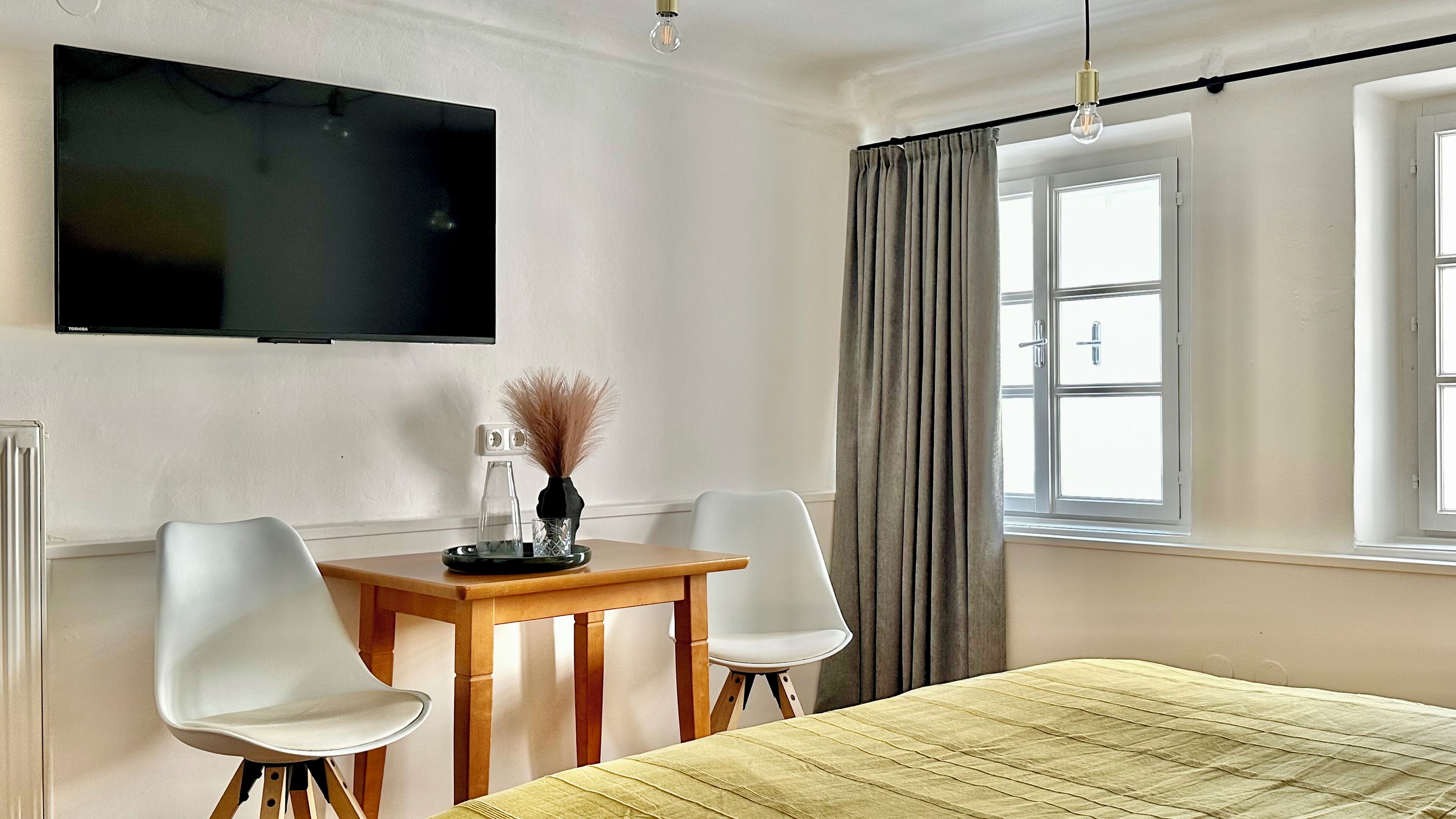 A hotel room with a bed, a TV mounted on the wall, a table with chairs, and a window with curtains.