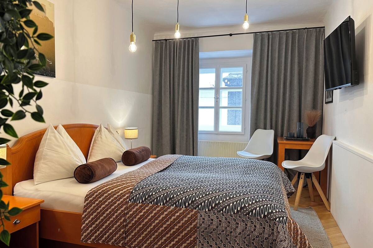A hotel room with a bed, bedside table, lamp, chair, desk, TV, and a window with curtains.