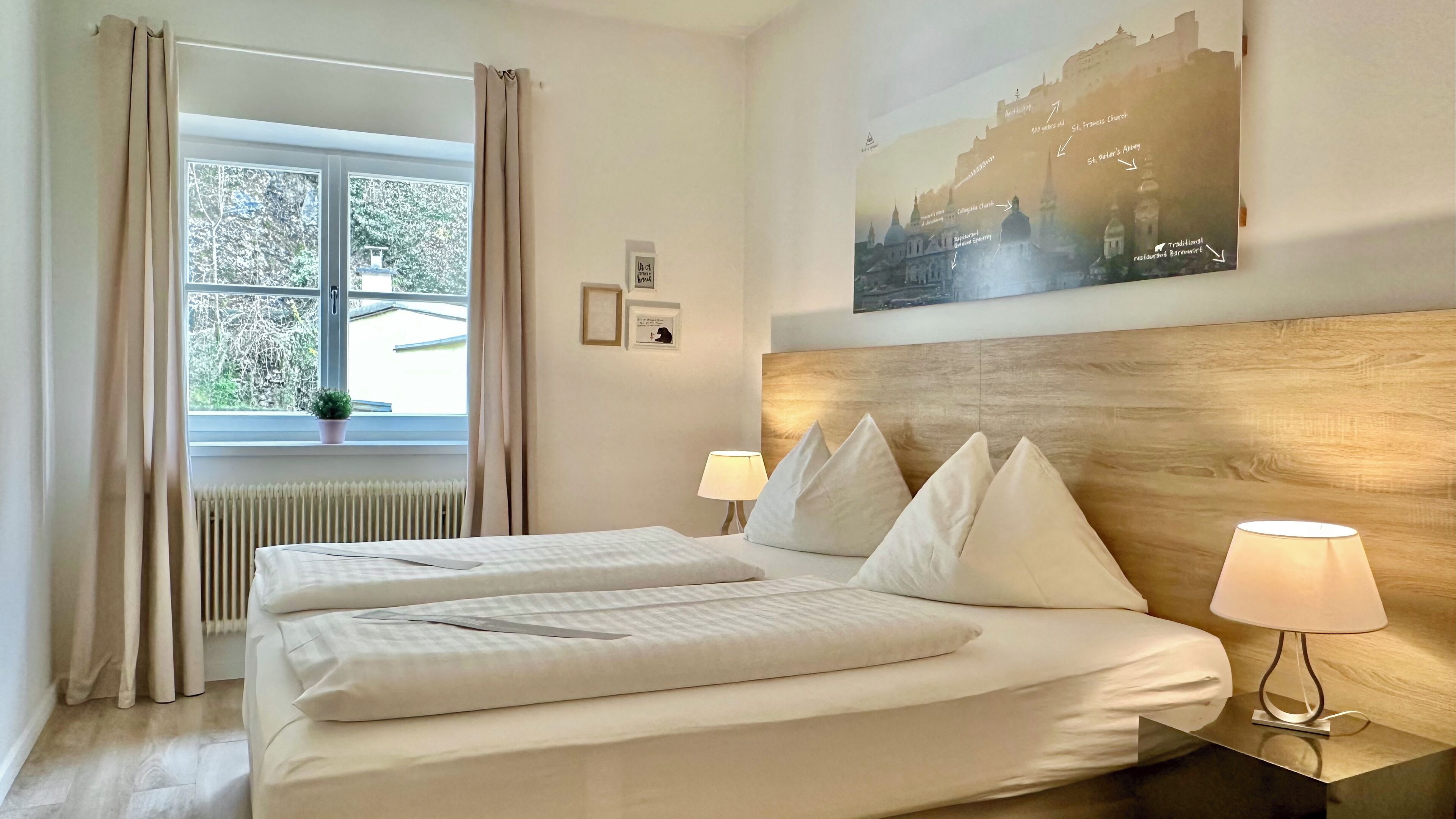A hotel room with a bed, two bedside lamps, a window with curtains, a radiator, and a wall-mounted painting.