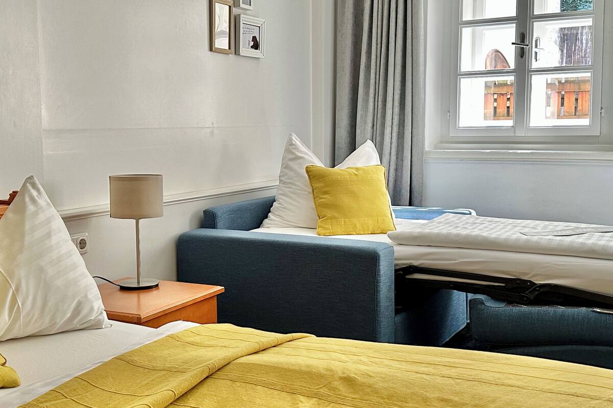 A hotel room with two beds, a bedside table with a lamp, and a window with a view of buildings.