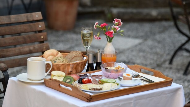 Ett frukostbricka med bröd, frukt, ost och drycker står på ett bord utomhus.