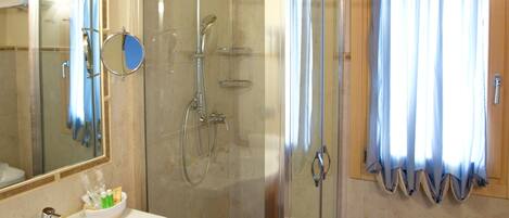 A bathroom with a glass-enclosed shower, a sink with a mirror, and a window with curtains.