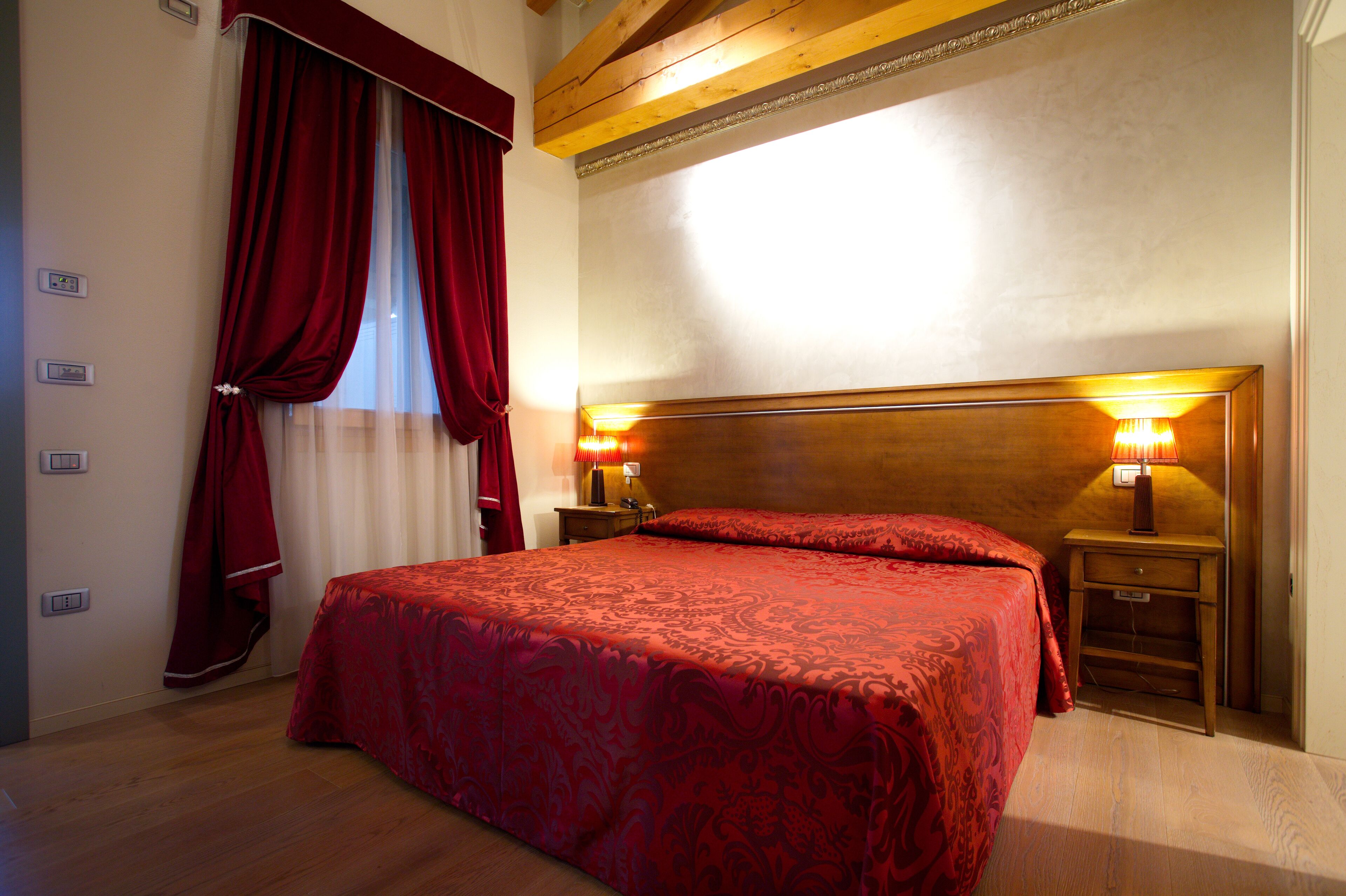 A hotel room with a large bed, wooden headboard, two bedside lamps, and a window with red curtains.
