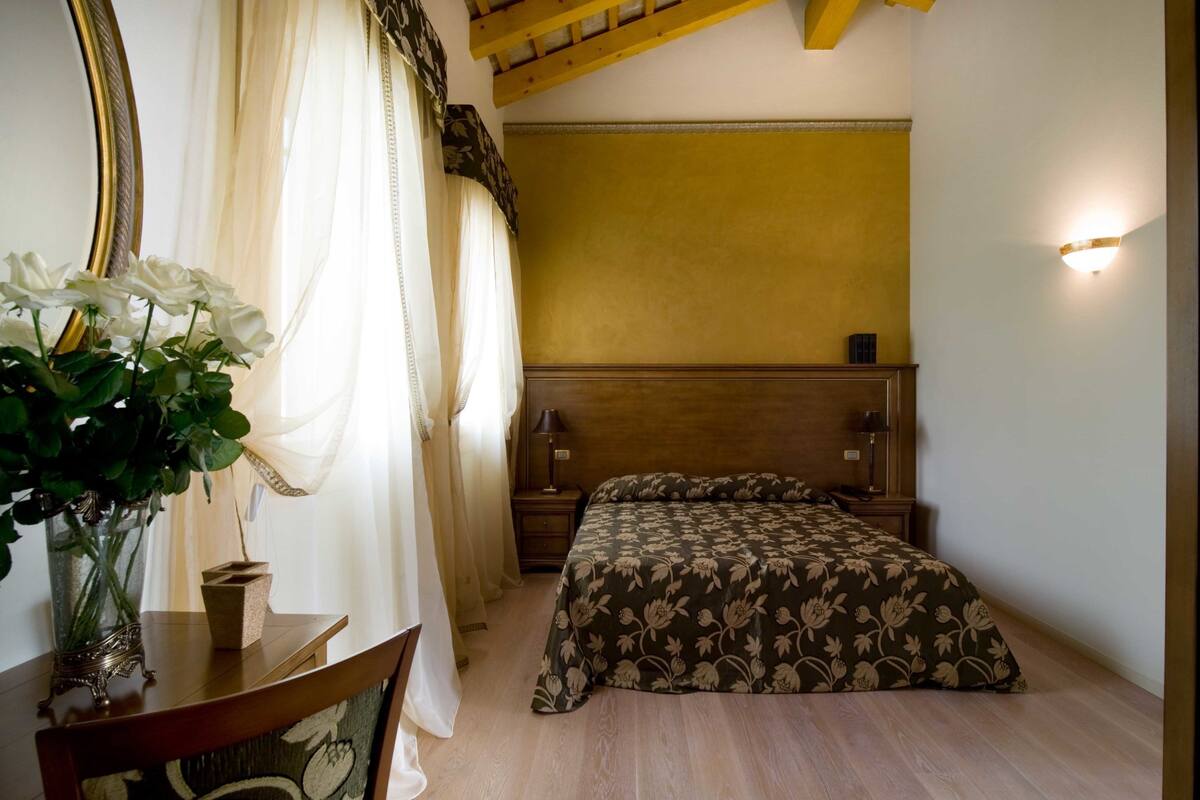 A bedroom with a wooden headboard, a floral bedspread, a bedside table with a lamp, and a vase with flowers on a wooden dresser.