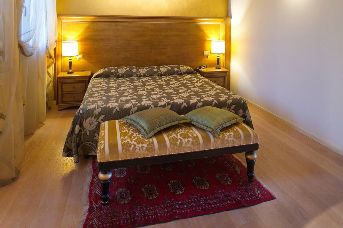 A bedroom with a wooden ceiling, a large bed with a patterned bedspread, two bedside lamps, and a red rug.