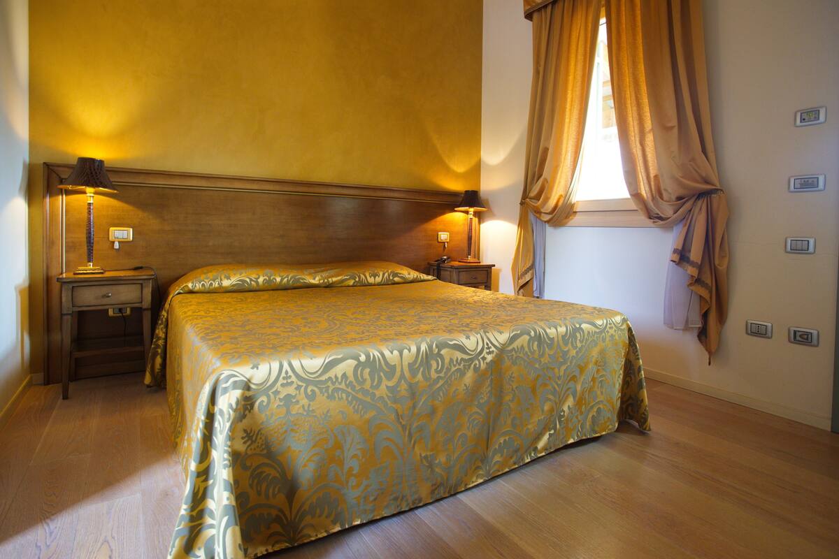 A hotel room with a large bed, two bedside tables with lamps, a window with curtains, and a wooden headboard.