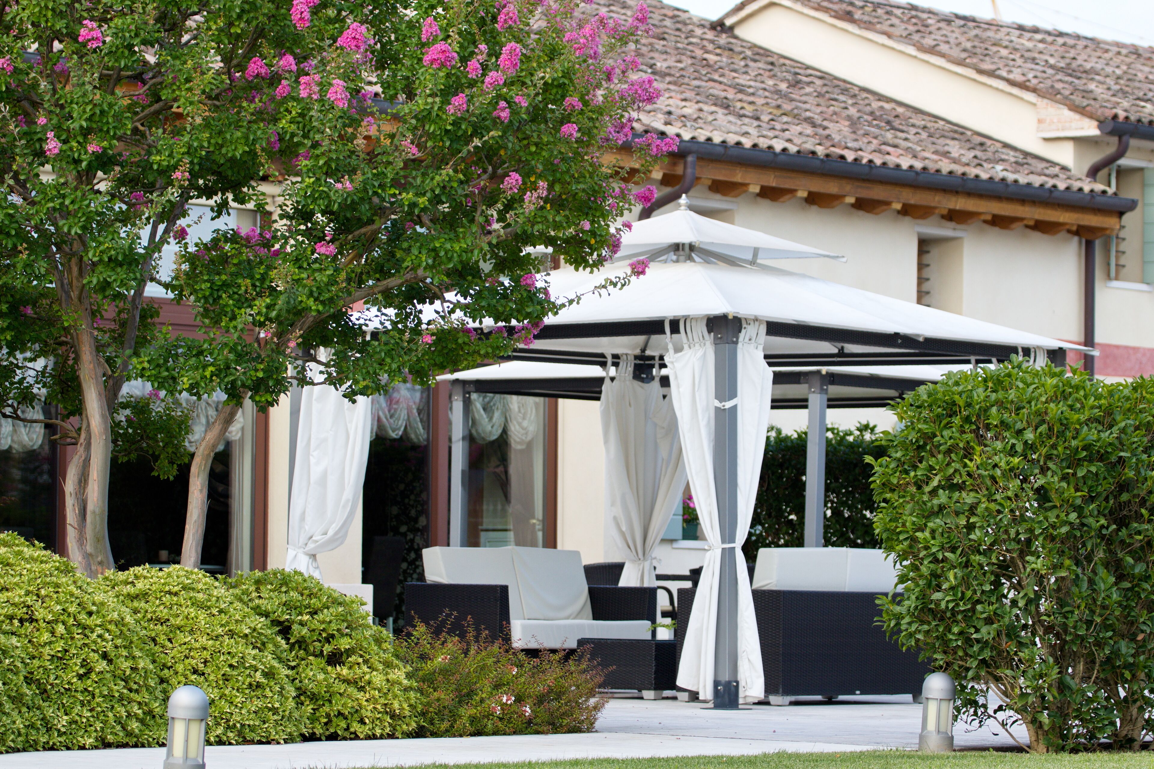 A covered outdoor seating area with white furniture, a pergola with white curtains, and a tree with pink flowers.