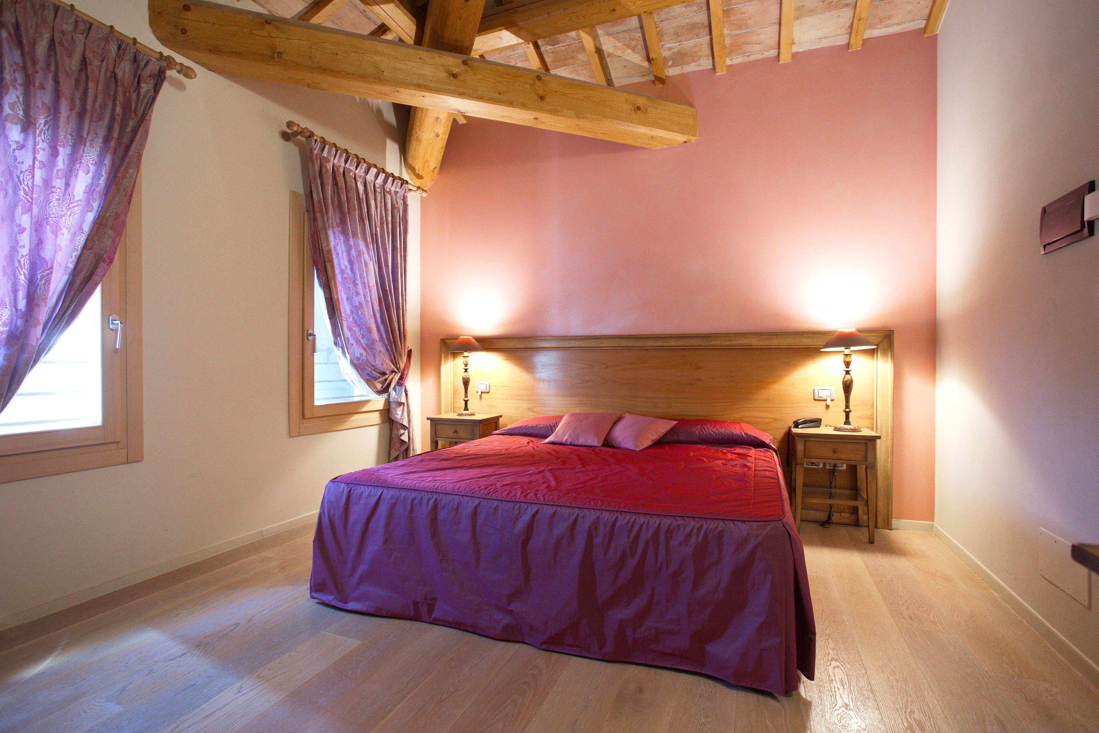Een slaapkamer met een houten hoofdbord, een bed met een rode sprei, twee nachtkastjes met lampen, en een raam met gordijnen.
