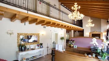 Una hall d'albergo con una reception, un grande specchio, un lampadario e un balcone con ringhiera in metallo.
