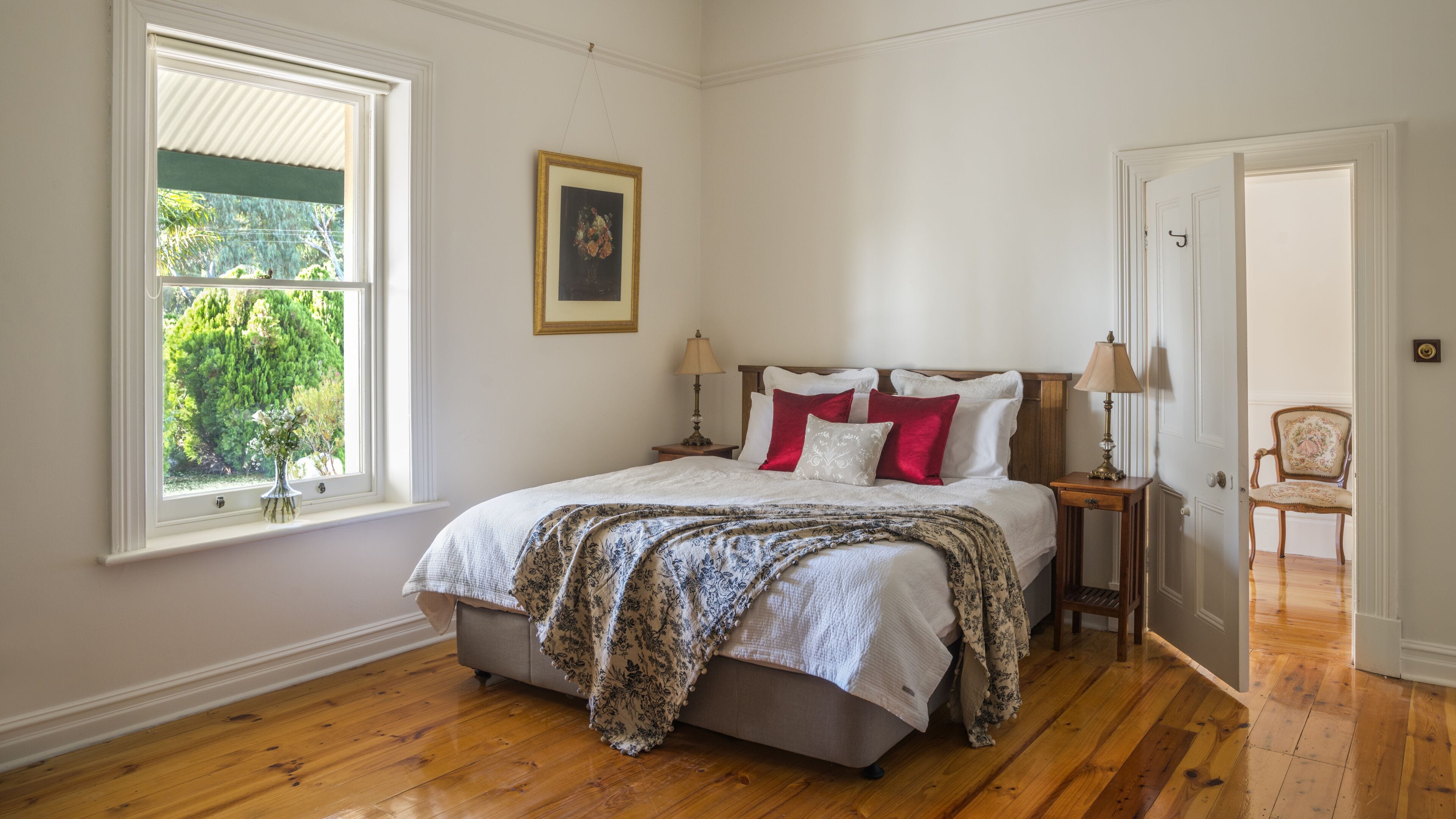 A bedroom with a large bed, a window with a view of greenery, a framed picture on the wall, and a chair in the corner.