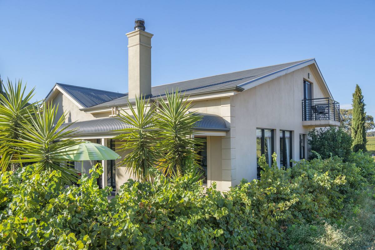 A modern two-story house with a prominent chimney, a balcony with a railing, and lush greenery in the foreground.