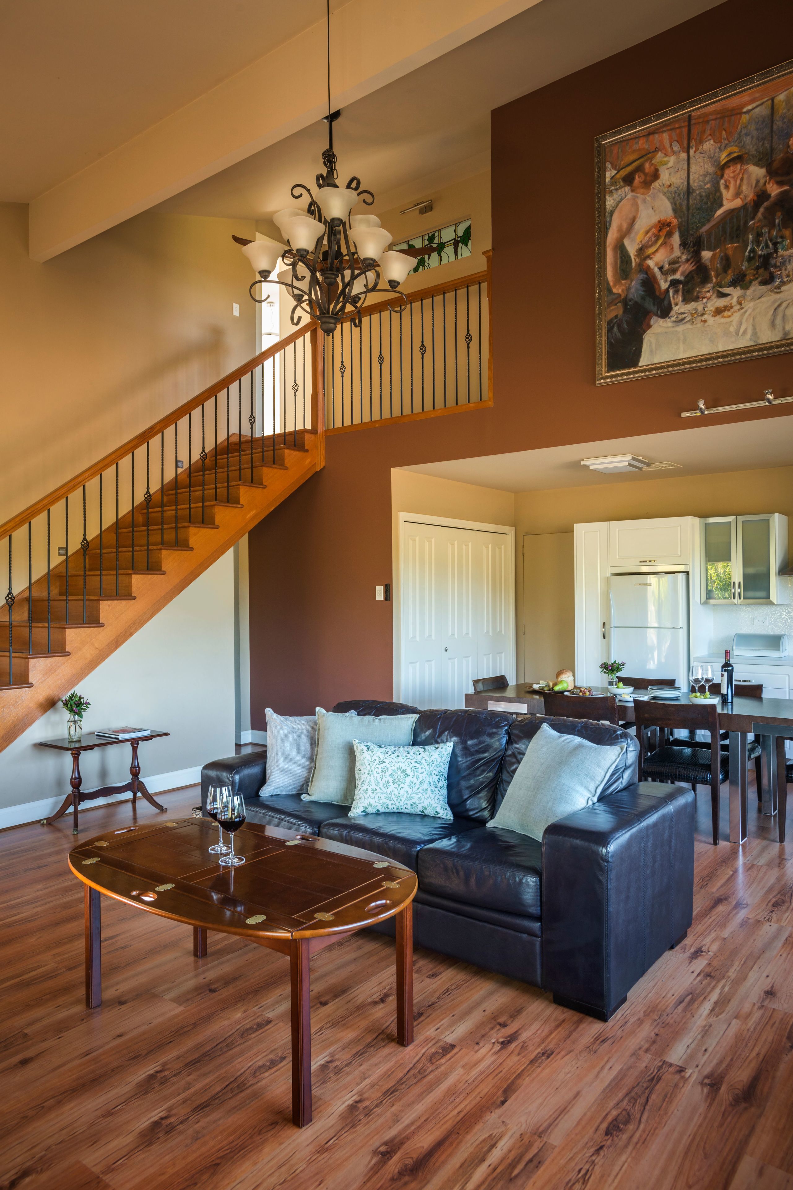 A spacious living room with a wooden staircase, a black leather sofa, a wooden coffee table, and a dining area with a kitchen in the background.