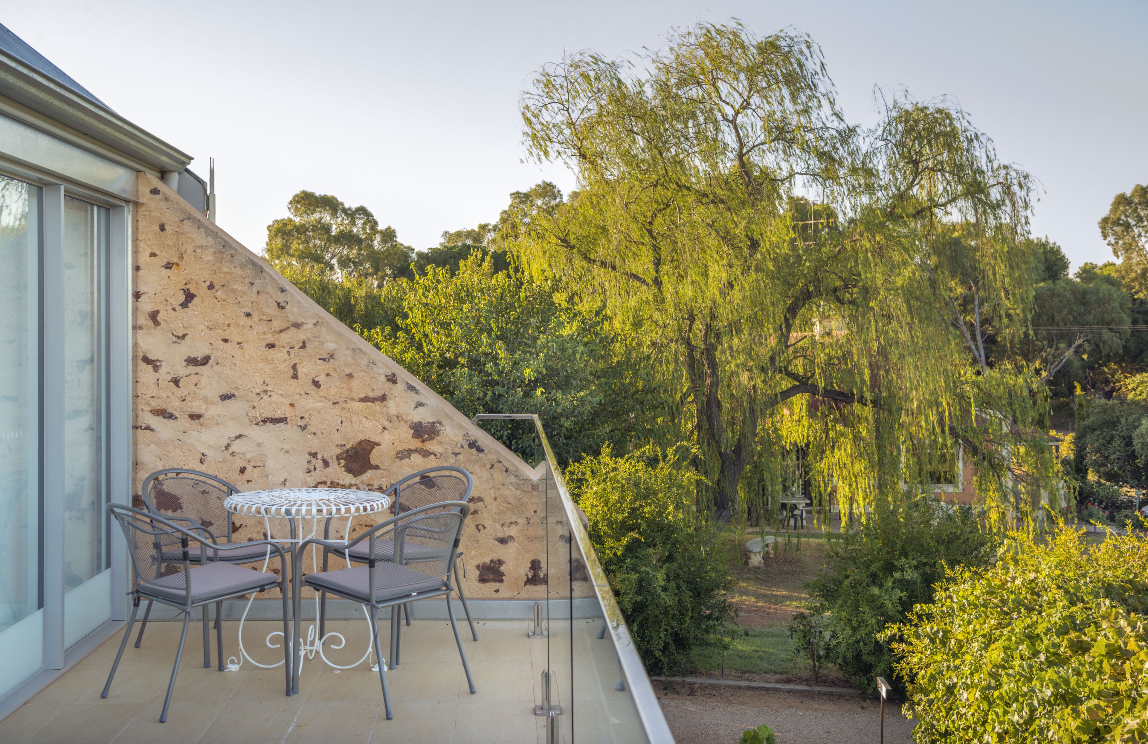 A balcony with a table and chairs, overlooking a garden with trees and shrubs.
