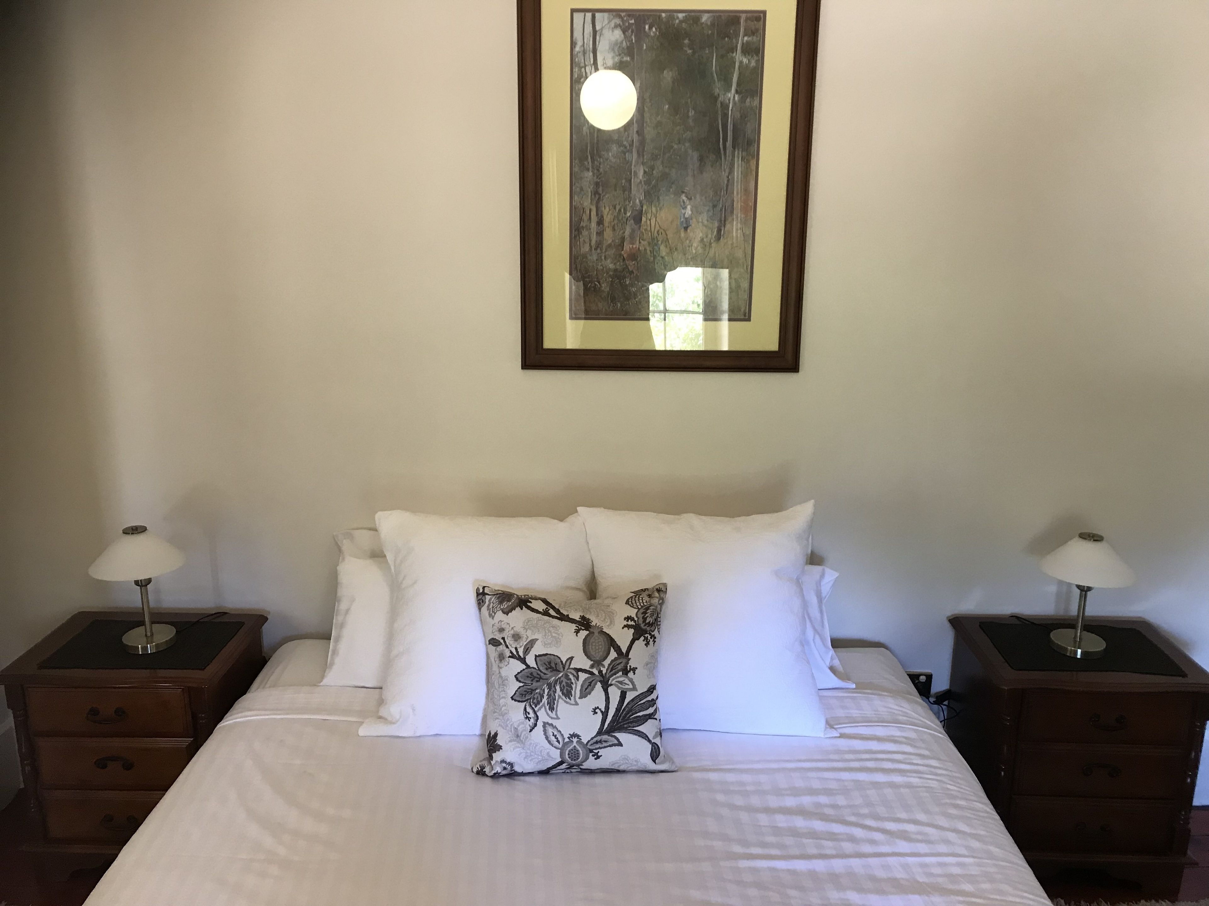 Una cama bien hecha con sábanas blancas, dos mesitas de noche con lámparas y una obra enmarcada en la pared.