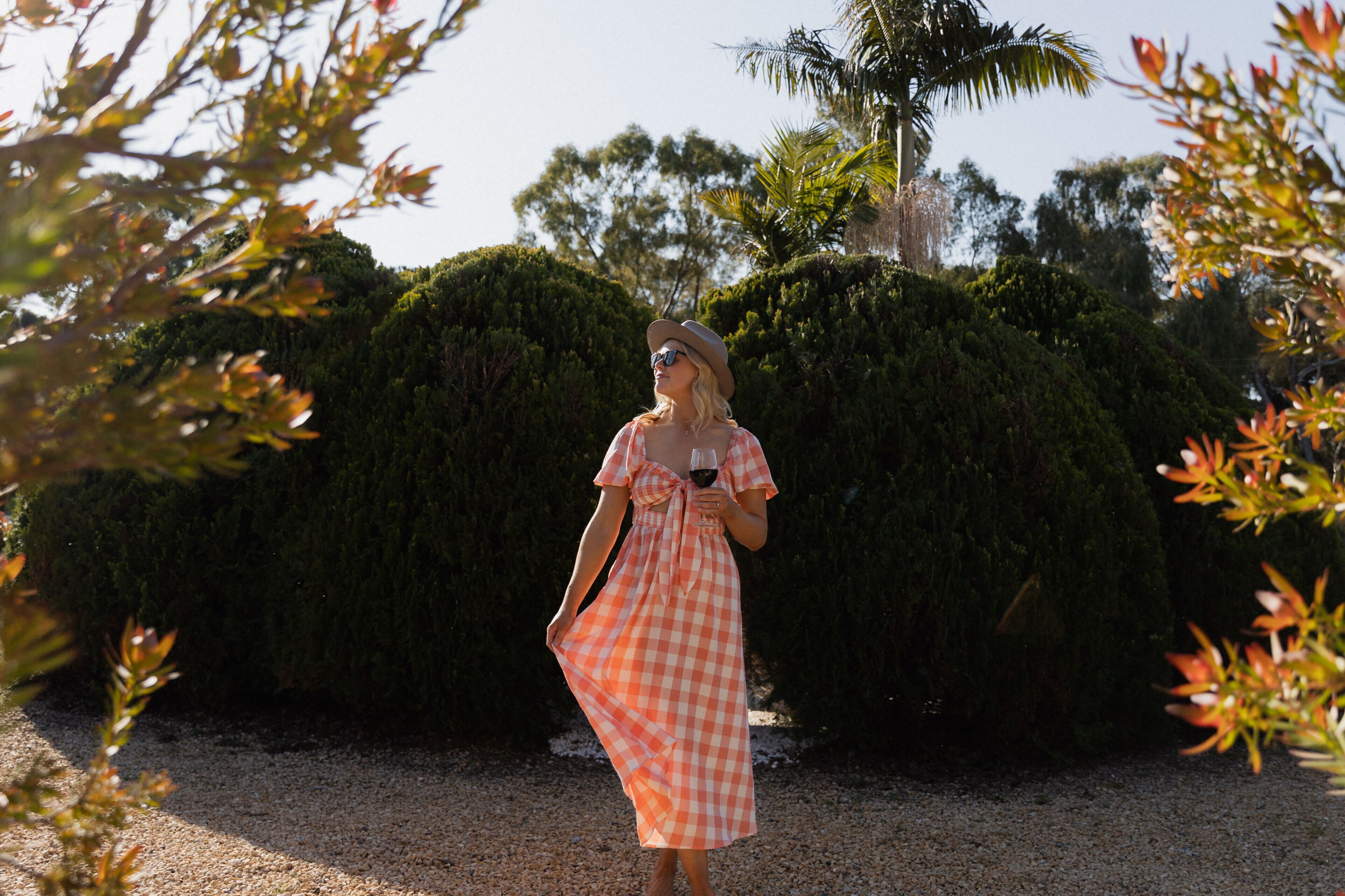 A woman in a checkered dress standing in a garden with a gravel path and neatly trimmed hedges.