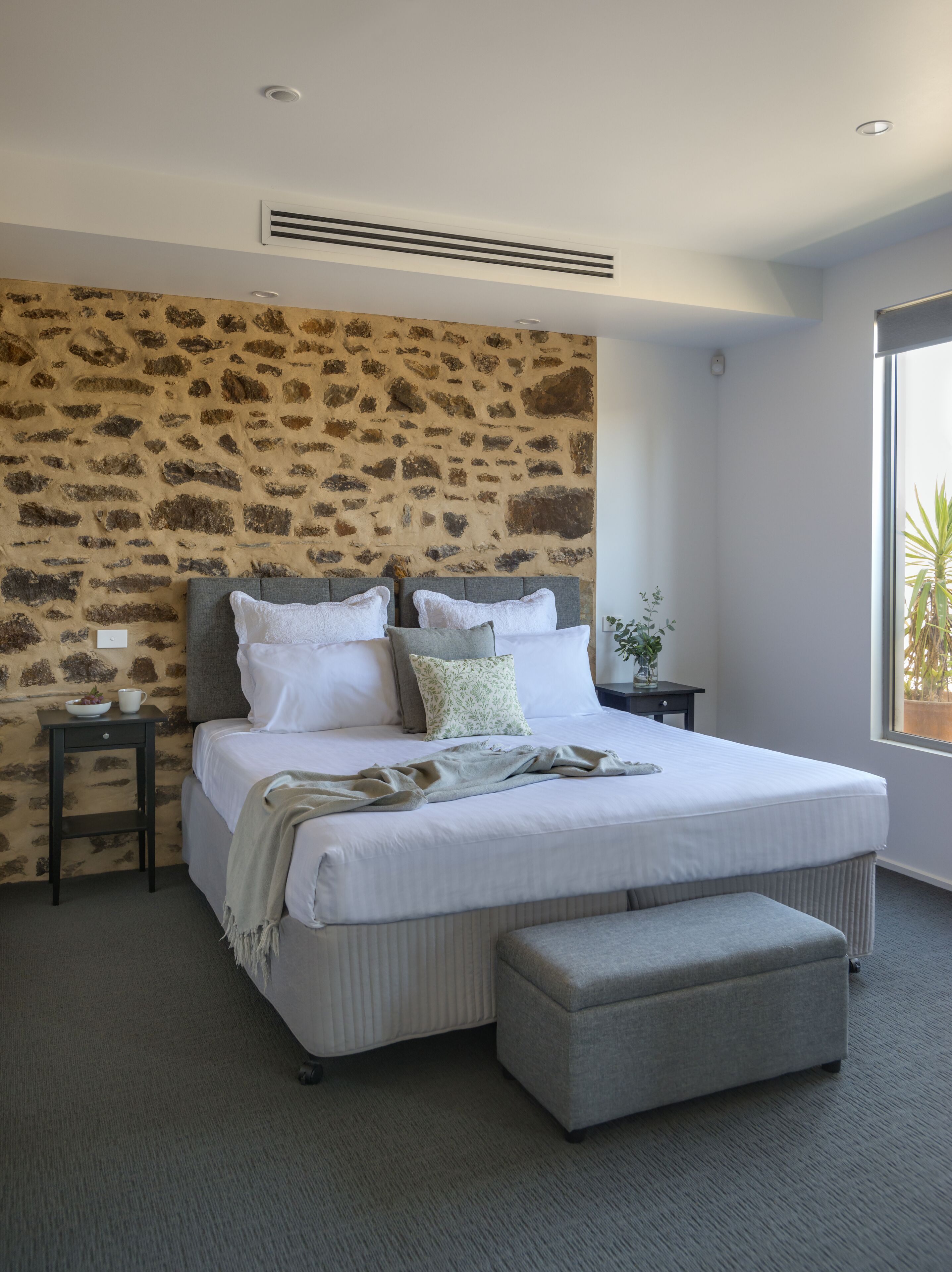 A bedroom with a stone-patterned headboard, a bed with white linens, a grey bench, and a nightstand with a vase.