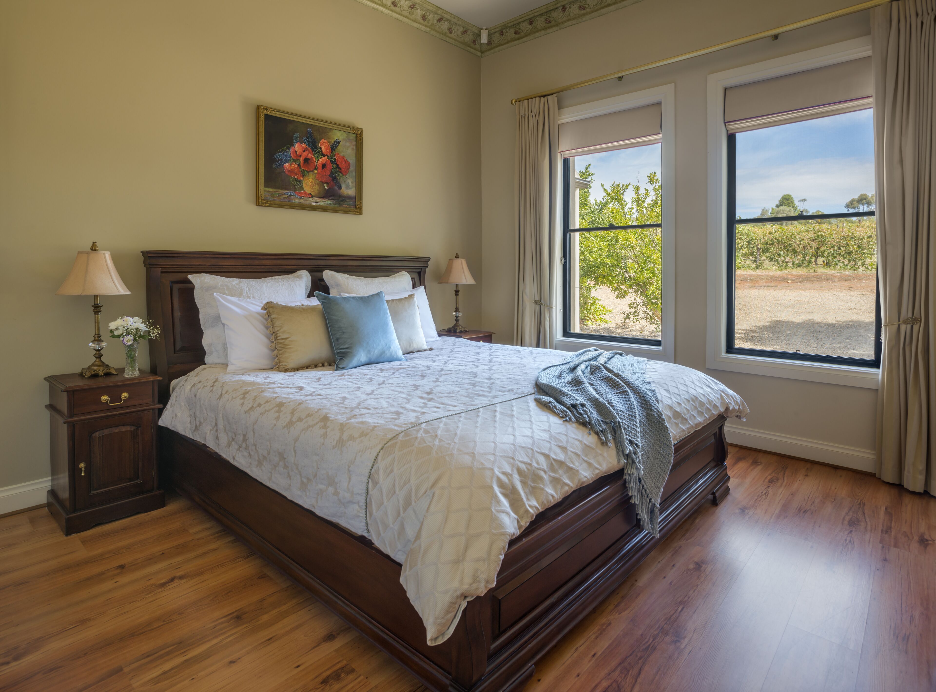 A neatly made bed with a quilt, bedside tables with lamps, a painting on the wall, and a window with a view of greenery.