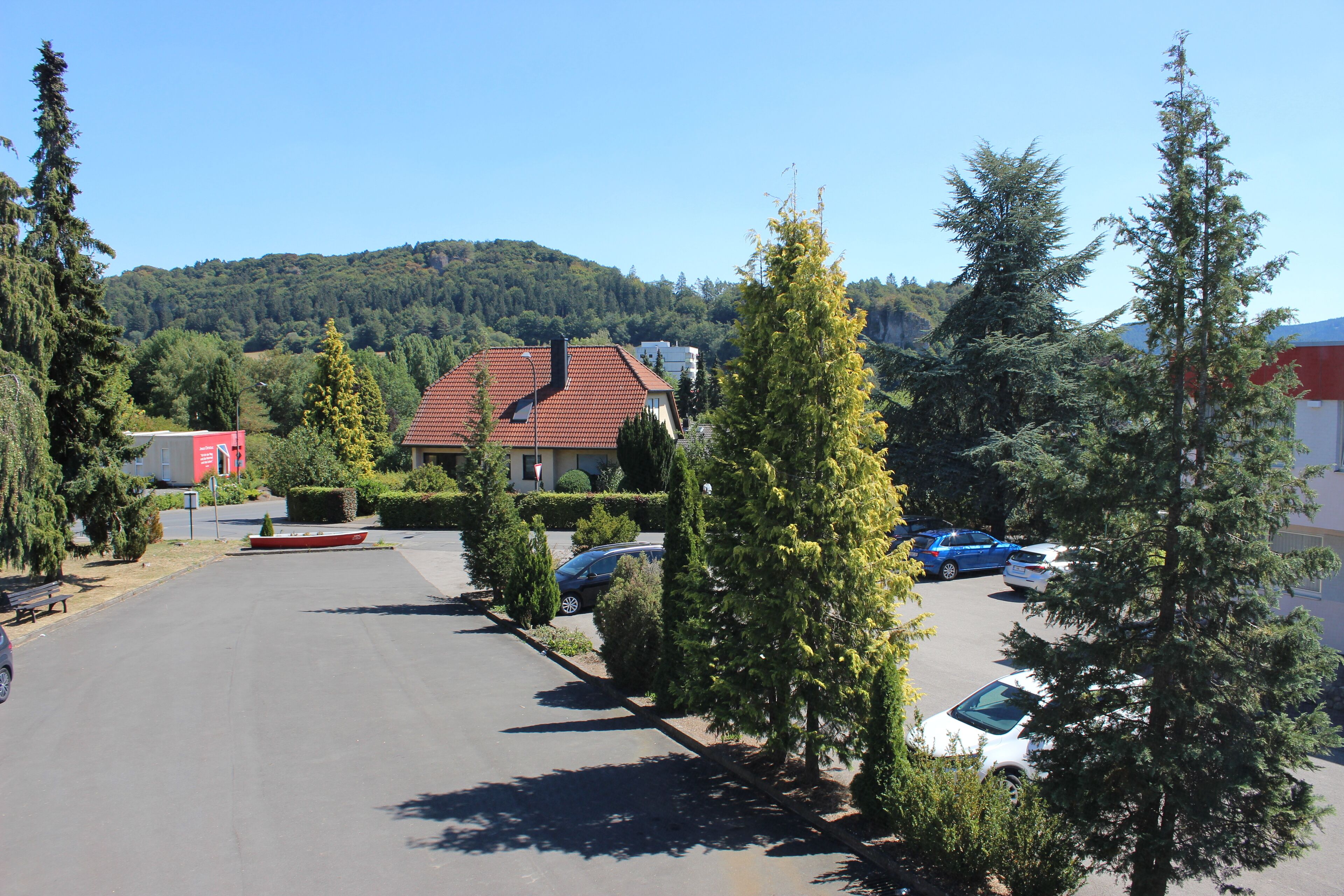 A parking lot with several cars, surrounded by trees and a building with a red roof.