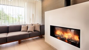 A modern living room with a built-in fireplace, a grey sofa with cushions, and a large window with sheer curtains. - Seehotel am Stausee (Gerolstein)