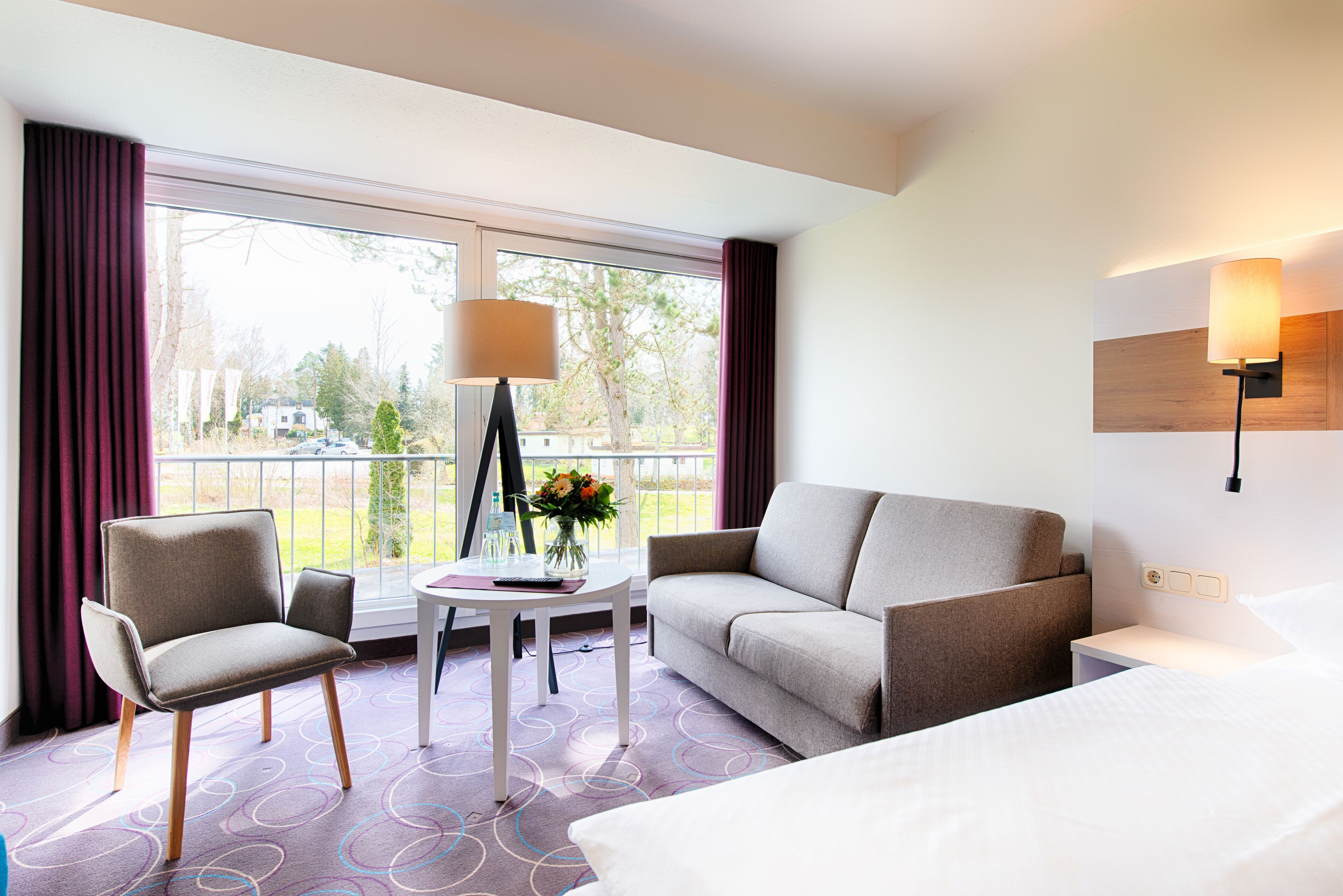 A hotel room with a bed, a sofa, a chair, a small table with a vase of flowers, and a lamp.