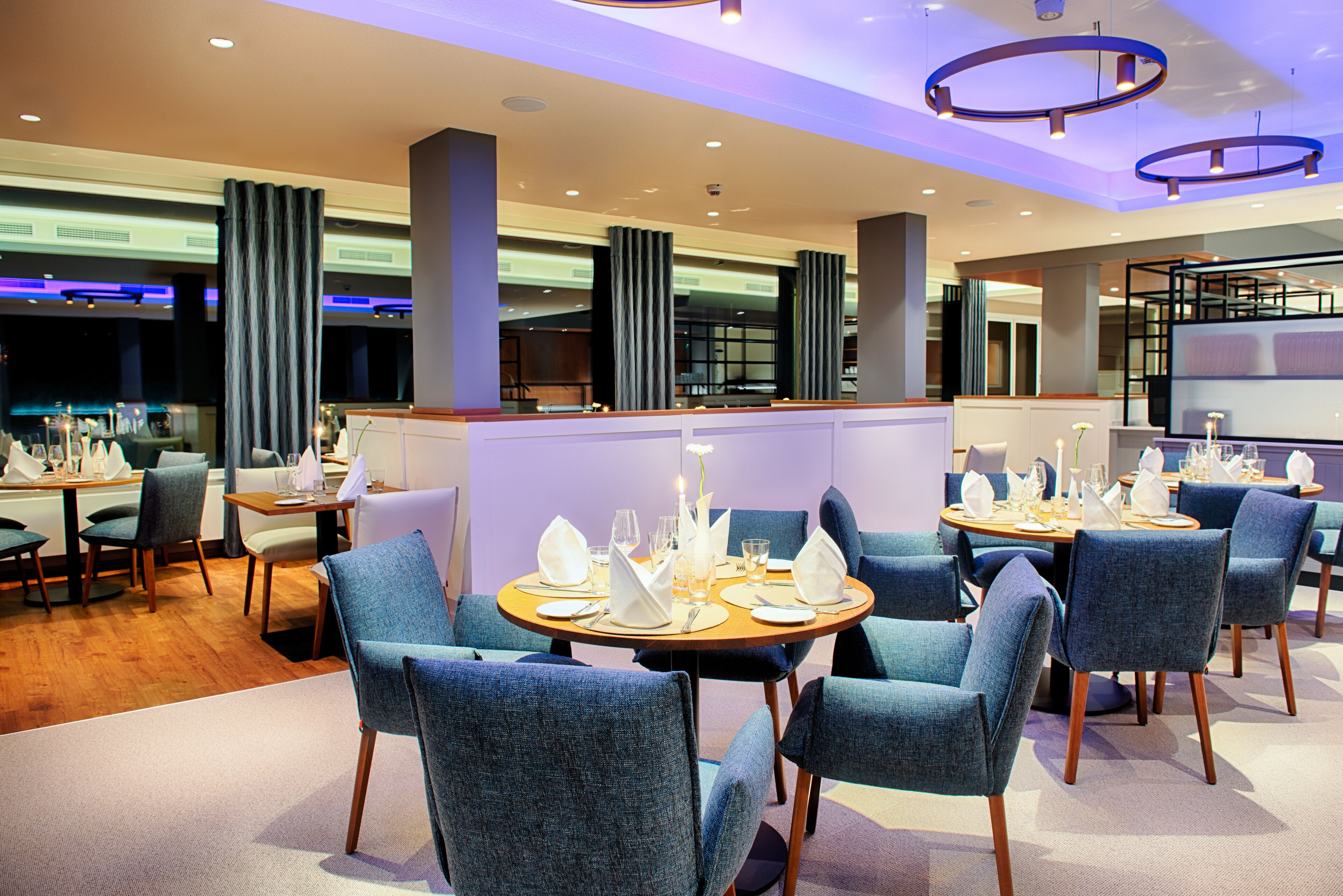 A modern restaurant with round tables, blue upholstered chairs, and a warm wooden floor.