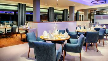 A modern restaurant with round tables, blue upholstered chairs, and a warm wooden floor.