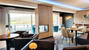 A modern hotel room with a fireplace, seating area, and large windows overlooking a scenic landscape. - Seehotel am Stausee (Gerolstein)